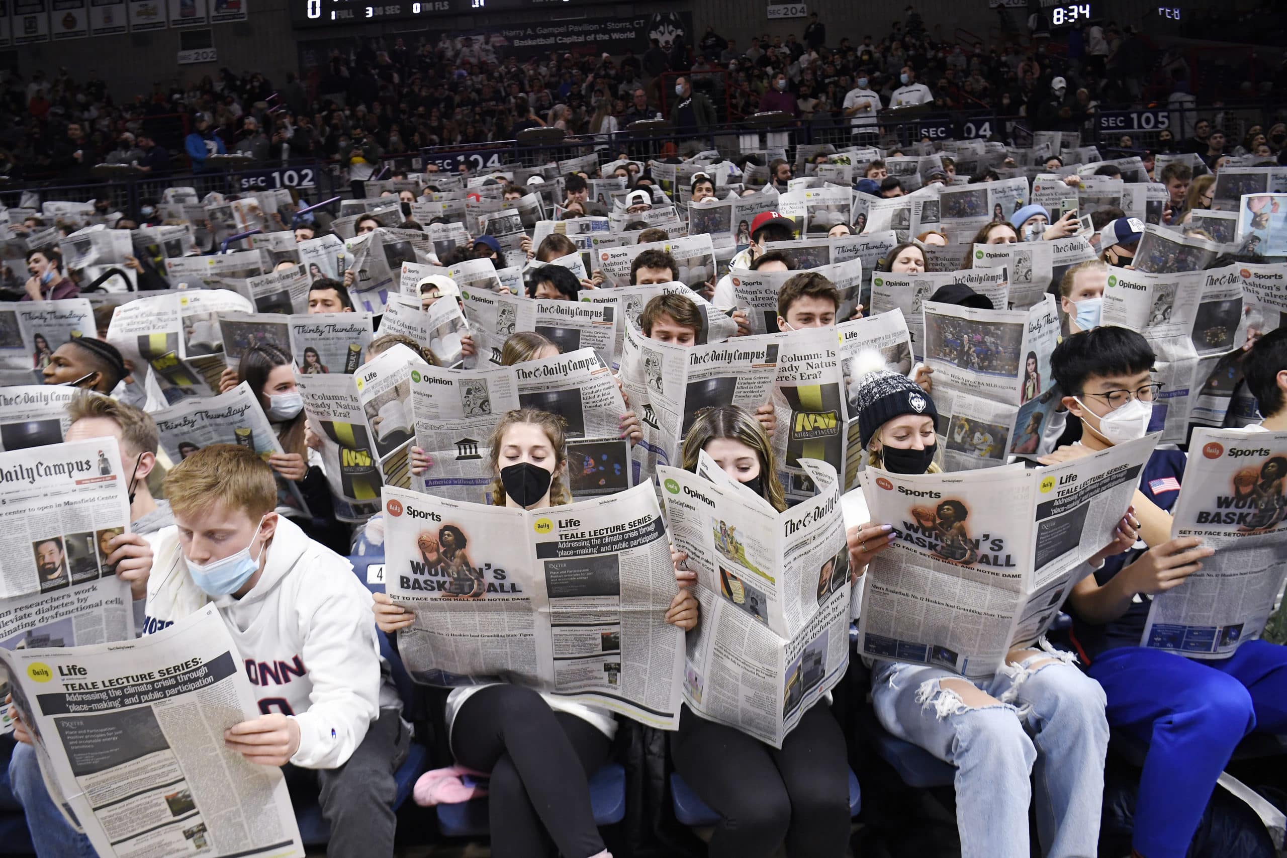 A crowd of students wearing face masks hold up newspapers in a basketball arena.
