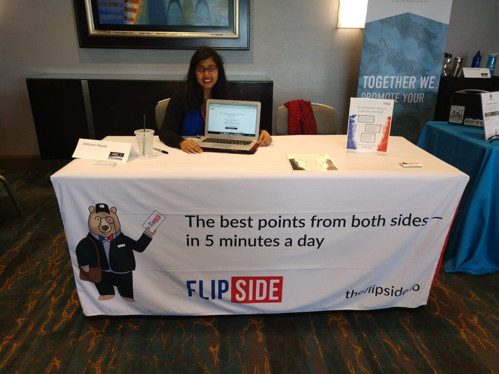 Annafi Wahead sits smiling at a table with a Flip Side banner. She holds a laptop computer with the Flip Side website pulled up.